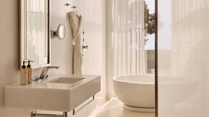 Light-filled bathroom with stone basin and freestanding tub, framed by sheer curtains and outdoor views.