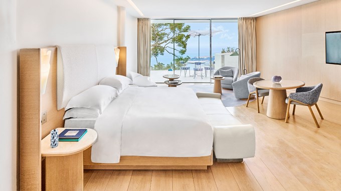 Spacious junior suite with a king bed, lounge and dining area in natural tones, opening onto a private terrace with panoramic views of the Mediterranean Sea and coastal landscape.