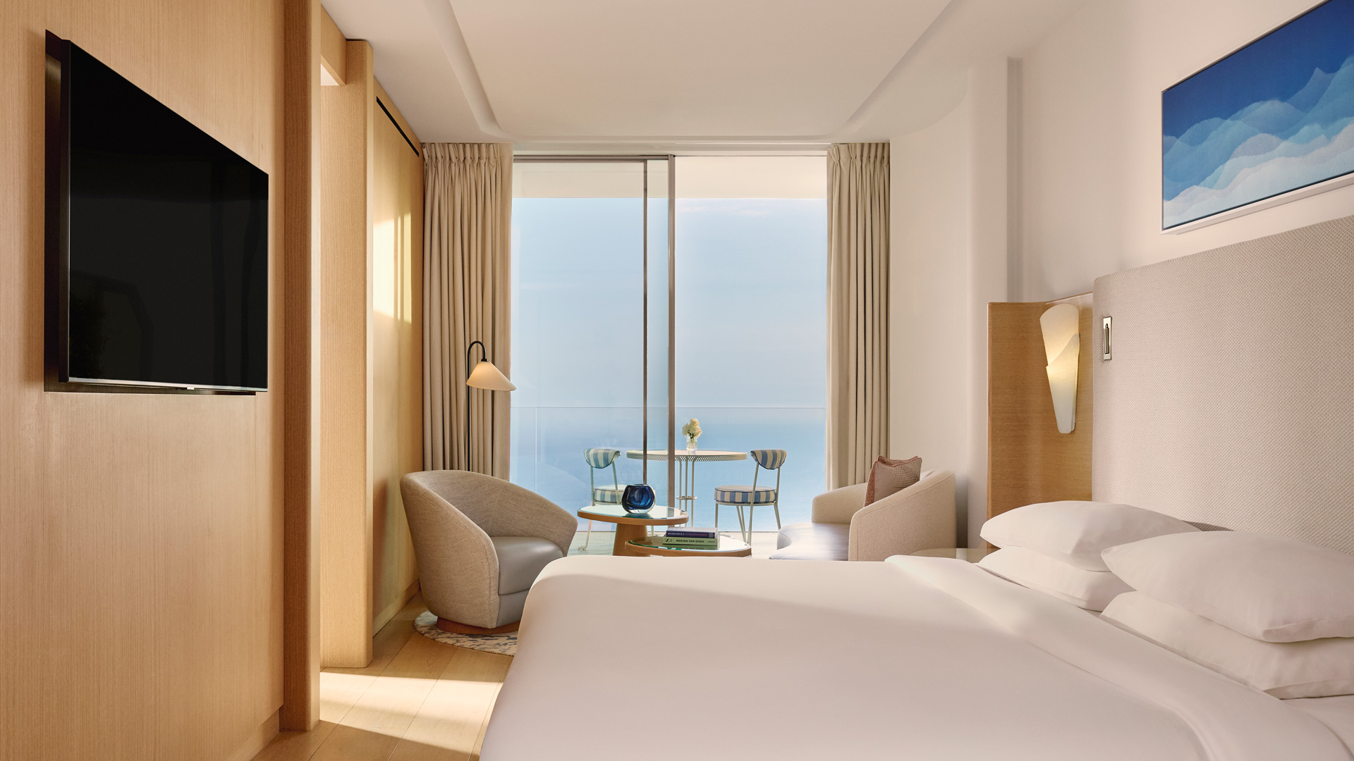 Vue intérieure d’un coin salon élégant donnant sur une terrasse privée avec table et chaises rayées bleues, et une vue dégagée sur la mer.