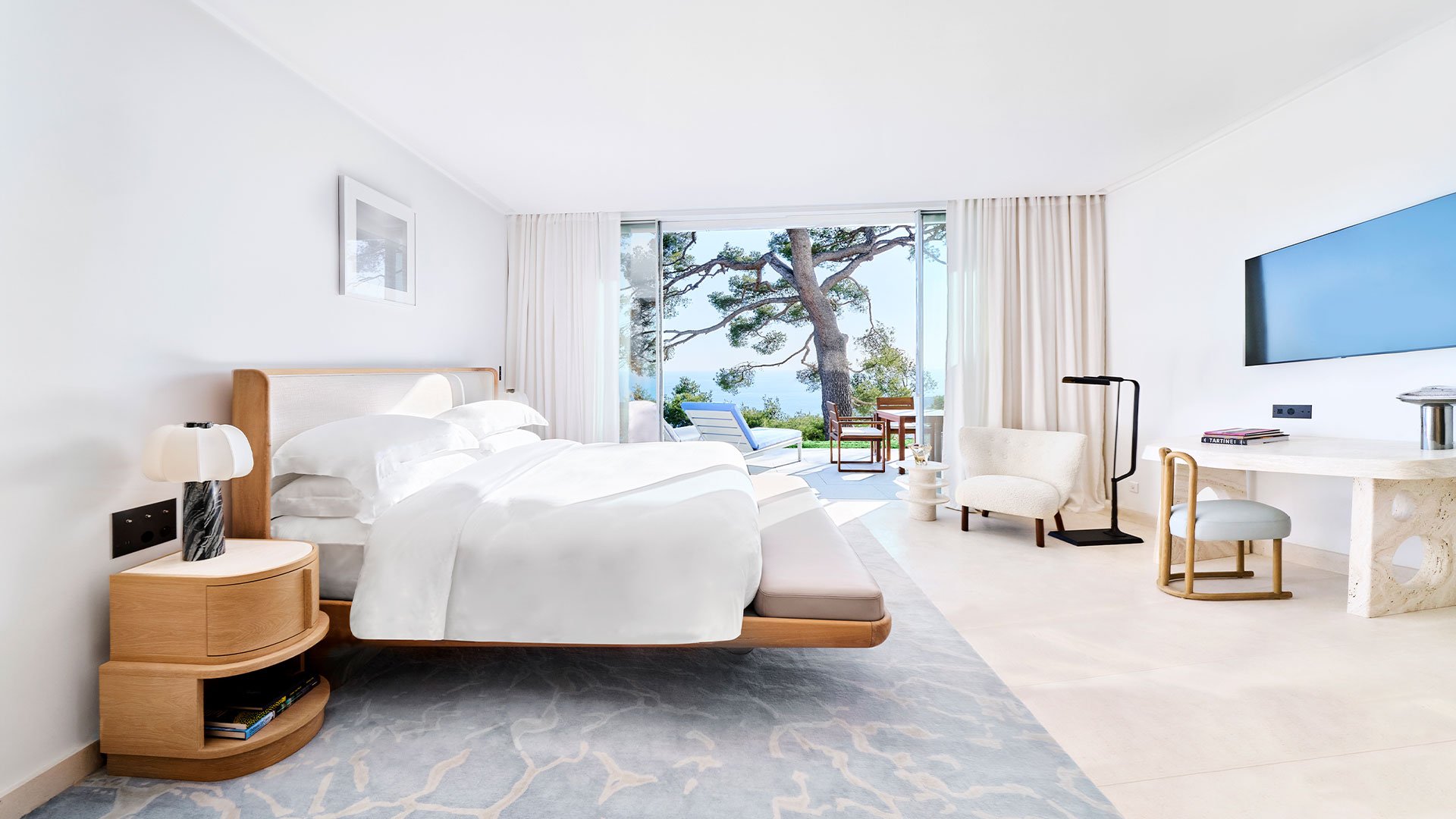 Studio grand luxe baigné de lumière naturelle, avec lit king-size, espace bureau et coin salon élégant, s’ouvrant sur une terrasse privée avec vue sur la mer Méditerranée et les pins de la Riviera.