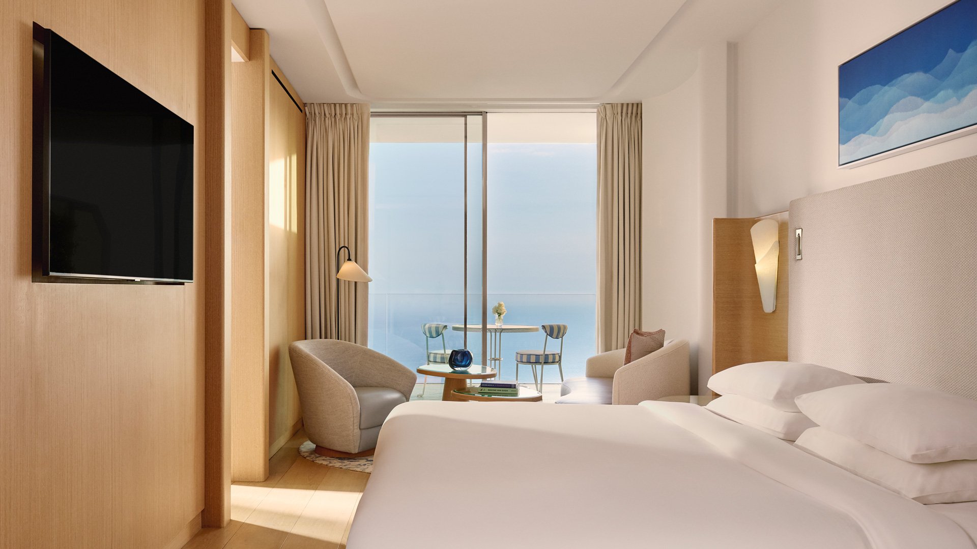 Contemporary hotel room with bed, armchairs, wall-mounted TV, and glass doors opening to sea view.