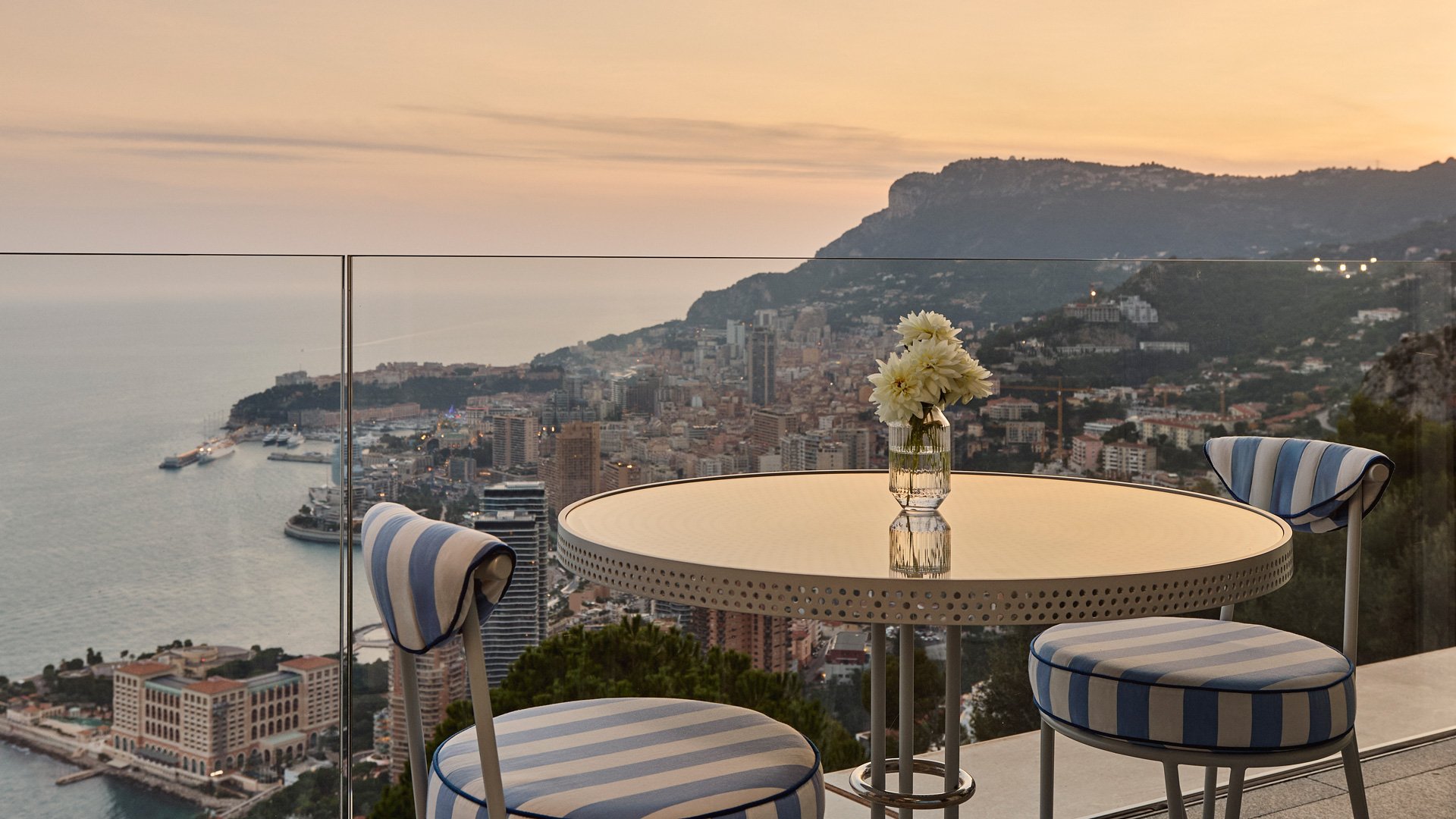 Balcony with striped chairs and round table, overlooking coastal cityscape at sunset.