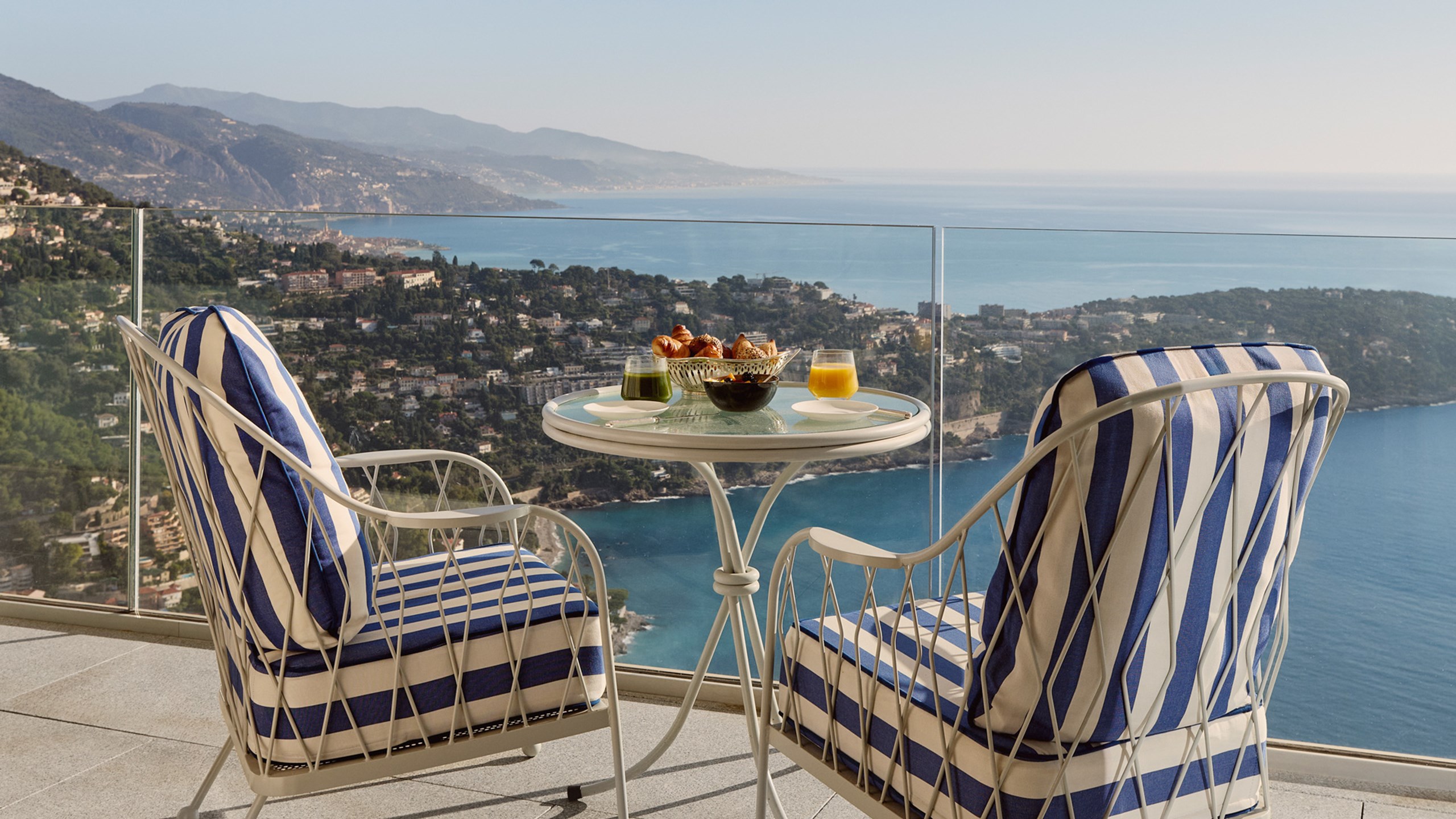 Table ronde en métal avec deux fauteuils rayés bleu et blanc sur une terrasse, offrant une vue spectaculaire sur la côte et les collines verdoyantes.