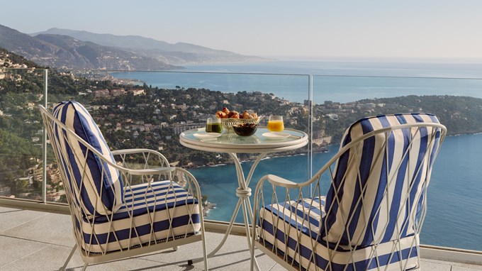 Table ronde en métal avec deux fauteuils rayés bleu et blanc sur une terrasse, offrant une vue spectaculaire sur la côte et les collines verdoyantes.
