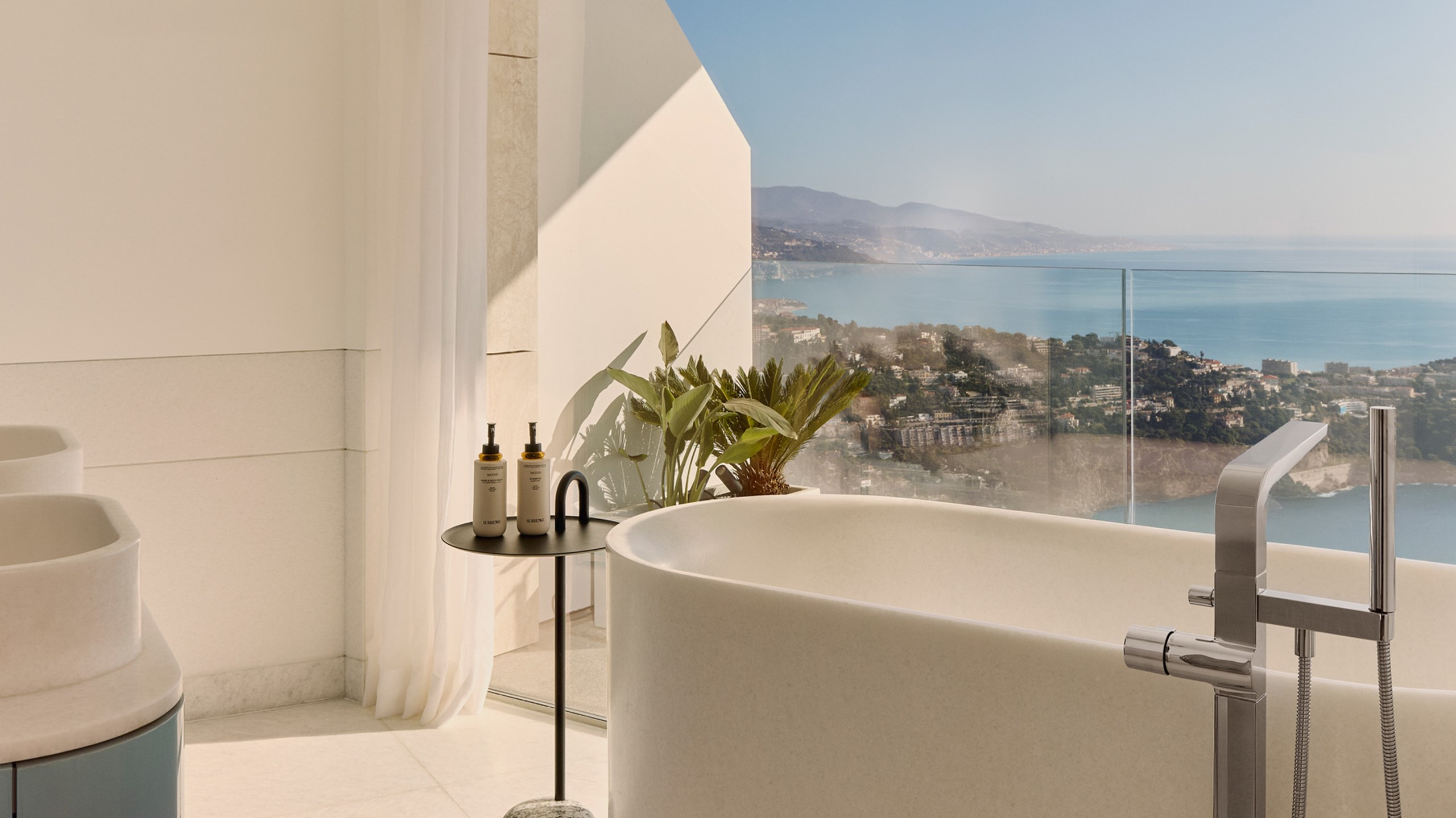 Salle de bain luxueuse avec baignoire îlot en pierre claire, grande fenêtre vitrée avec vue imprenable sur la mer et la côte.