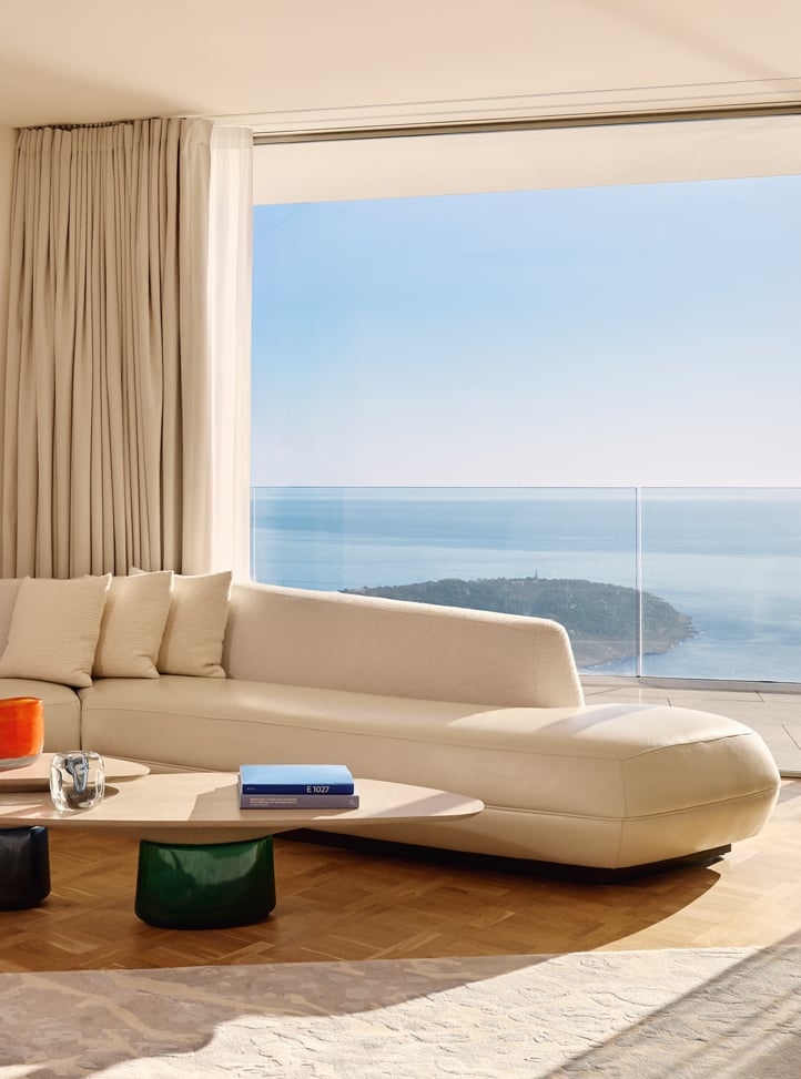 Un salon lumineux avec canapé en L couleur crème, table basse design et grande baie vitrée donnant sur la mer et un promontoire verdoyant.