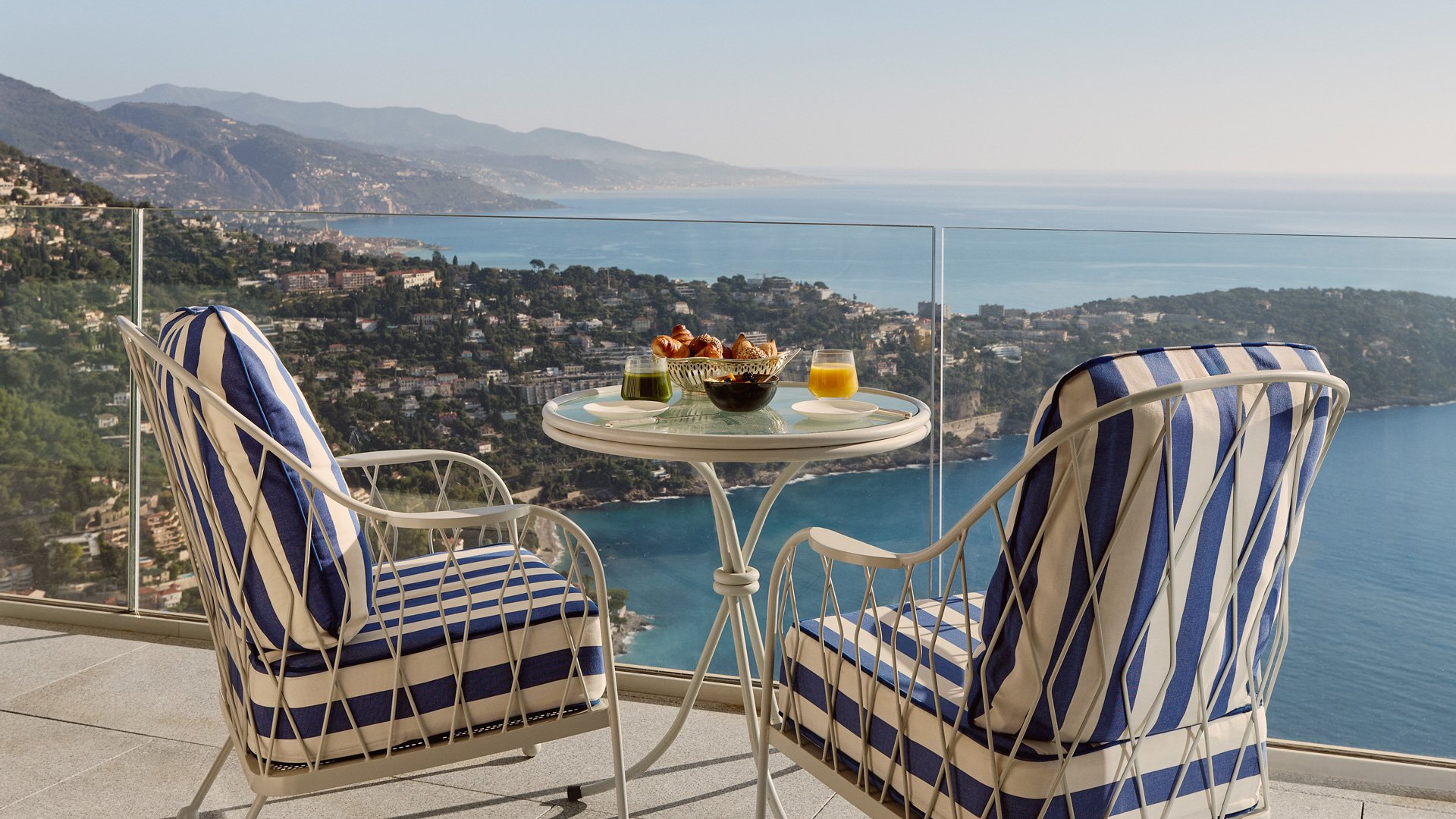 Glass-topped balcony table set for breakfast, overlooking the coastline and hillside homes.