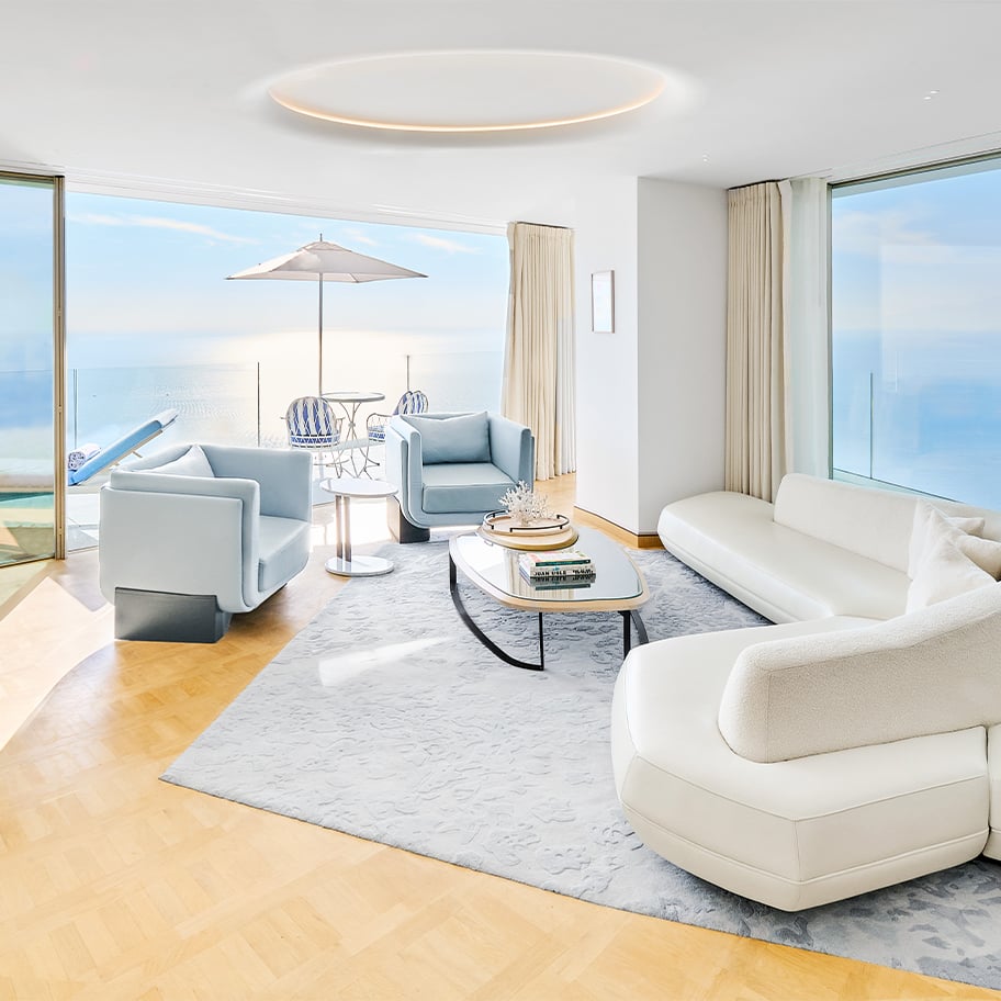 Spacious lounge with modern white furniture and large windows, opening to a terrace with sun loungers and sea horizon.