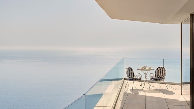 Terrasse privée avec deux chaises rayées et une table ronde surplombant une mer bleue paisible sous un ciel clair.