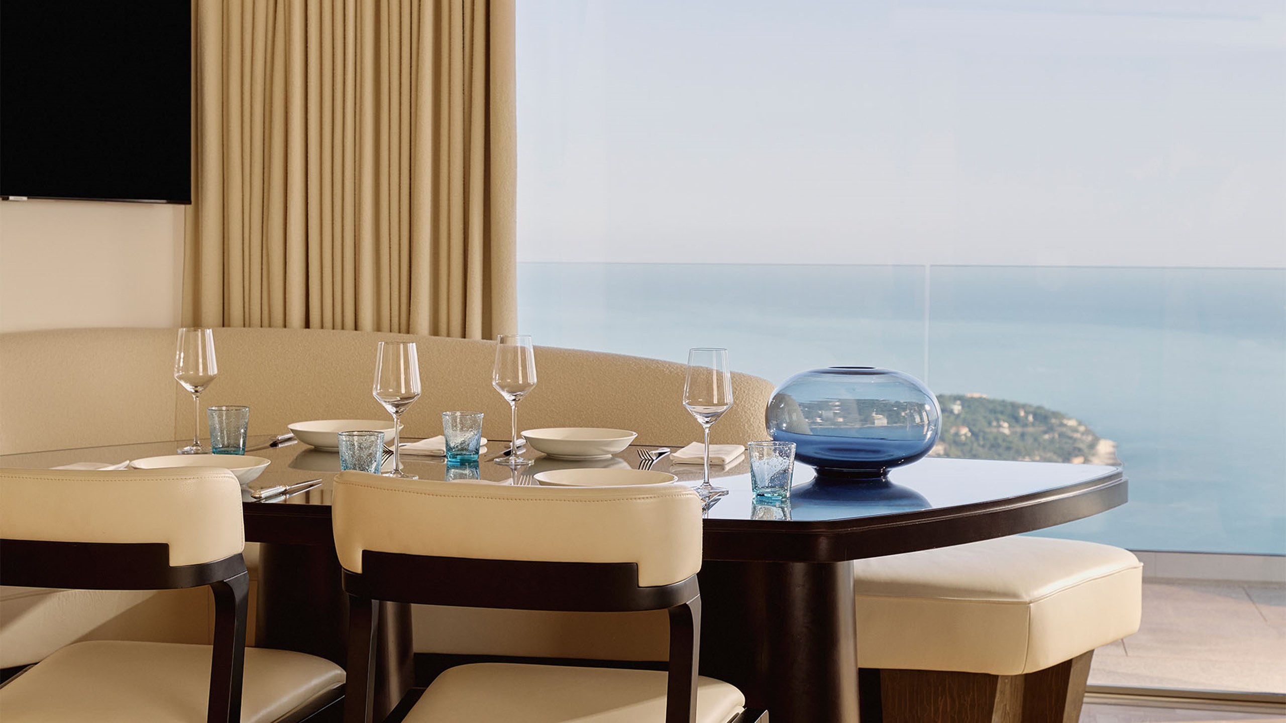 Chic dining area with a glass-topped table, cushioned chairs, blue-tinted glasses, and views across the coastline and sea.