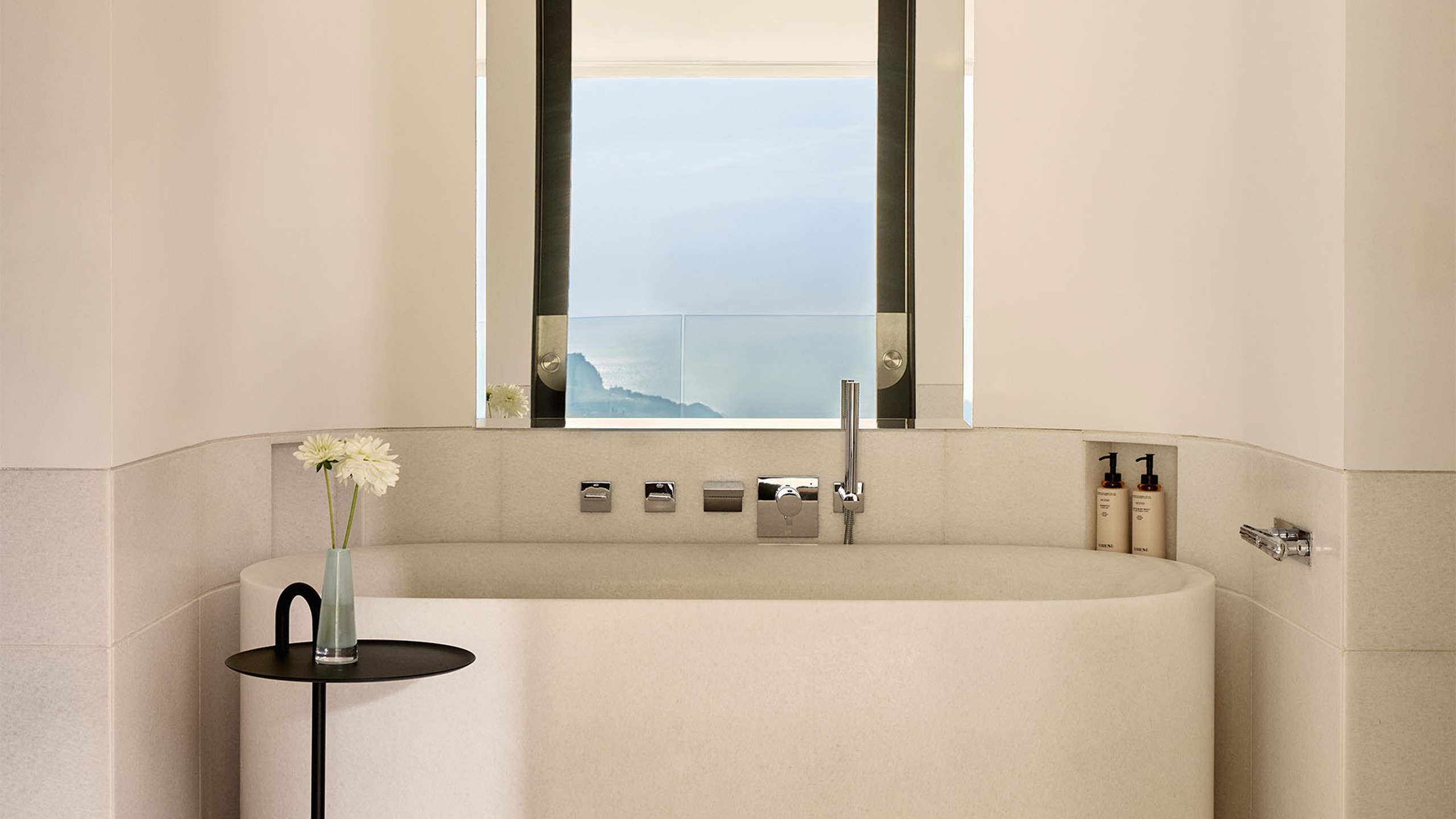 Minimalist bathroom with a freestanding tub, modern fixtures, and a window framing serene sea and mountain views.
