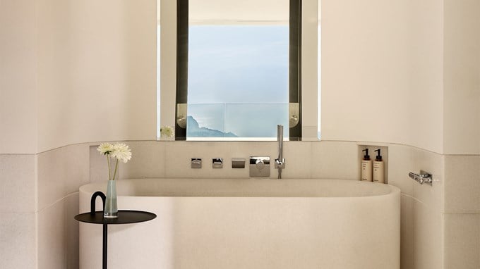 Minimalist bathroom with a freestanding tub, modern fixtures, and a window framing serene sea and mountain views.