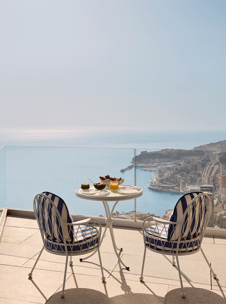 Elegant terrace setup with striped chairs and a breakfast table overlooking the Mediterranean coastline and harbour on a clear day.