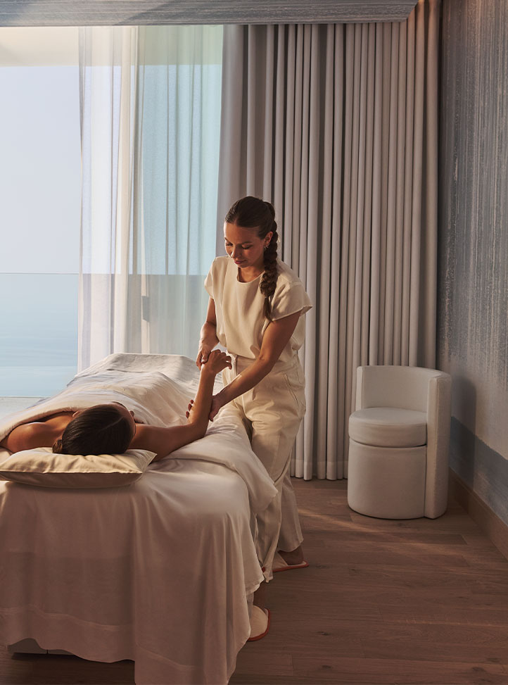 A spa therapist in neutral attire performs an arm massage on a client lying on a treatment bed in a softly lit room with ocean views through sheer curtains.
