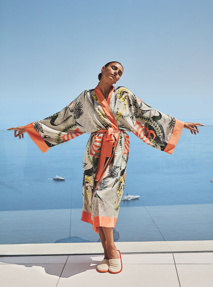 A woman in a printed silk robe with coral trim stands on a terrace overlooking a calm blue sea, arms outstretched and eyes closed in a relaxed pose.