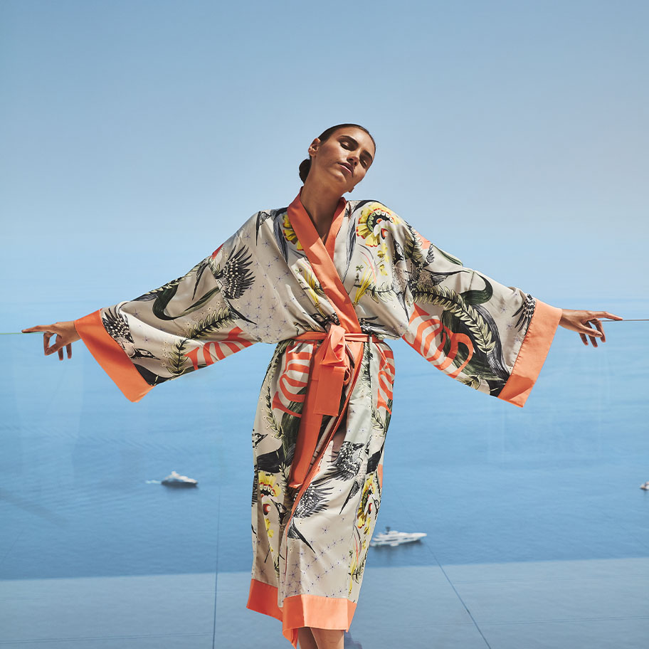 A woman in a patterned silk robe with coral trim stands on a terrace overlooking a calm blue sea, arms outstretched and eyes closed in a serene pose beneath a clear sky.