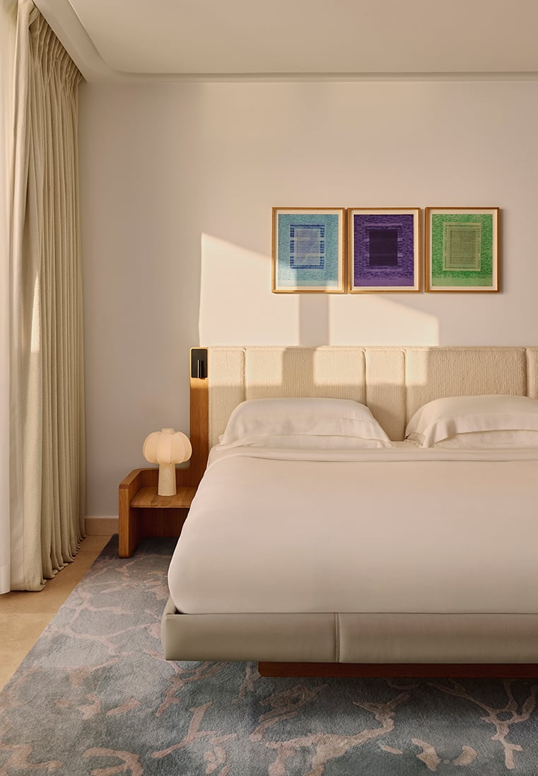Sunlit bedroom with a minimalist design, featuring a double bed with cream bedding, light wood headboard and matching bedside tables, a sculptural lamp on the left, and three colourful geometric artworks above the bed, set against a pale wall and light curtains.
