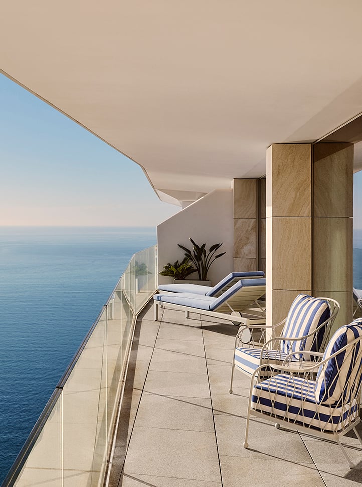 Private terrace with panoramic sea views, featuring two striped blue-and-white armchairs in the foreground and two cushioned sun loungers near the back, all set against a glass balustrade and light stone flooring, under a wide overhang.