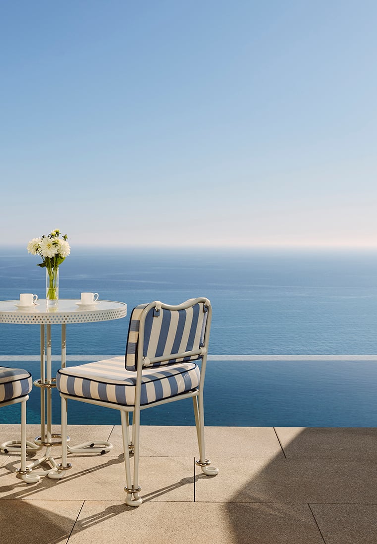 Outdoor seating area with a round white table and blue-and-white striped cushioned chairs, set on a stone terrace overlooking an infinity pool and the open sea under a clear blue sky, with a vase of white flowers and two coffee cups on the table.