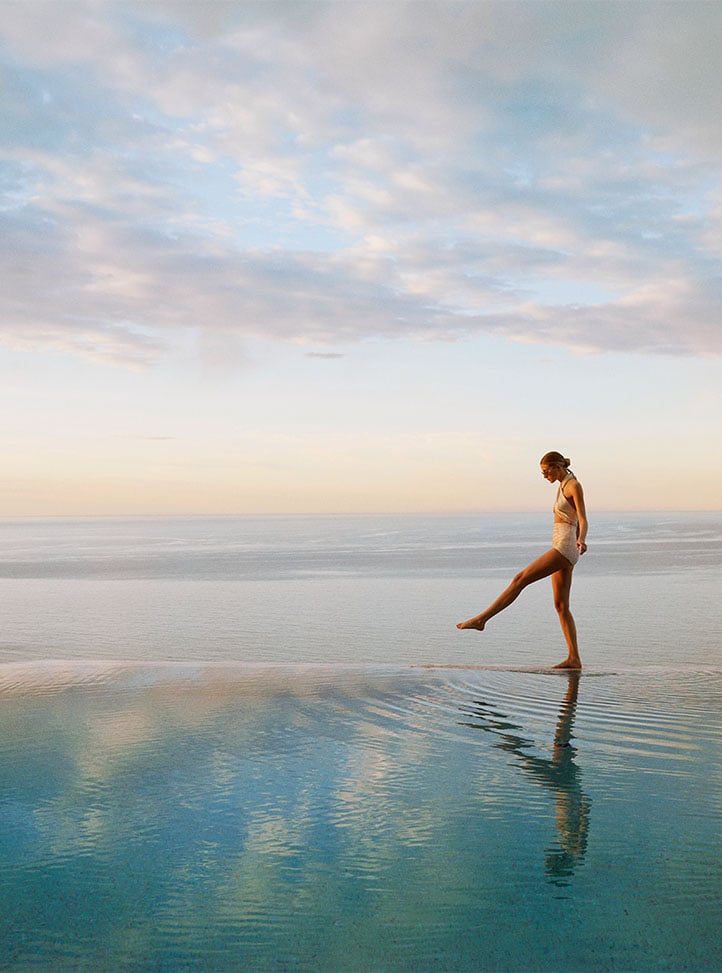 Woman in a swimsuit stepping into an infinity pool overlooking a calm sea at sunrise.