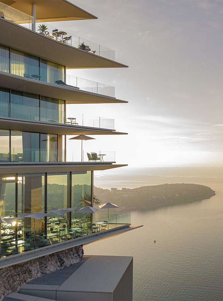 Modern clifftop hotel with tiered glass balconies overlooking the sea at sunset.