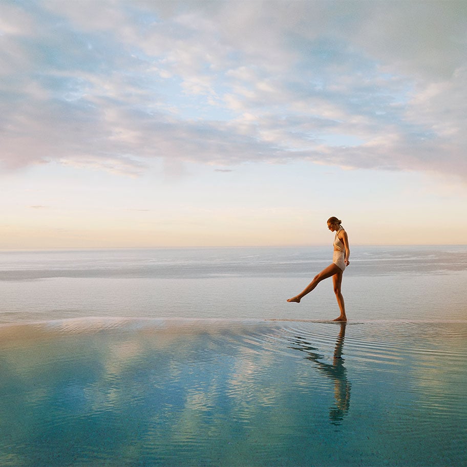 Woman in a swimsuit stepping into an infinity pool overlooking a calm sea at sunrise.