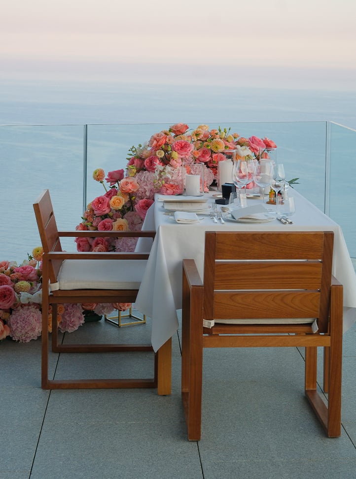 A romantic oceanfront table with elegant tableware, candles, and lush pink and peach florals.