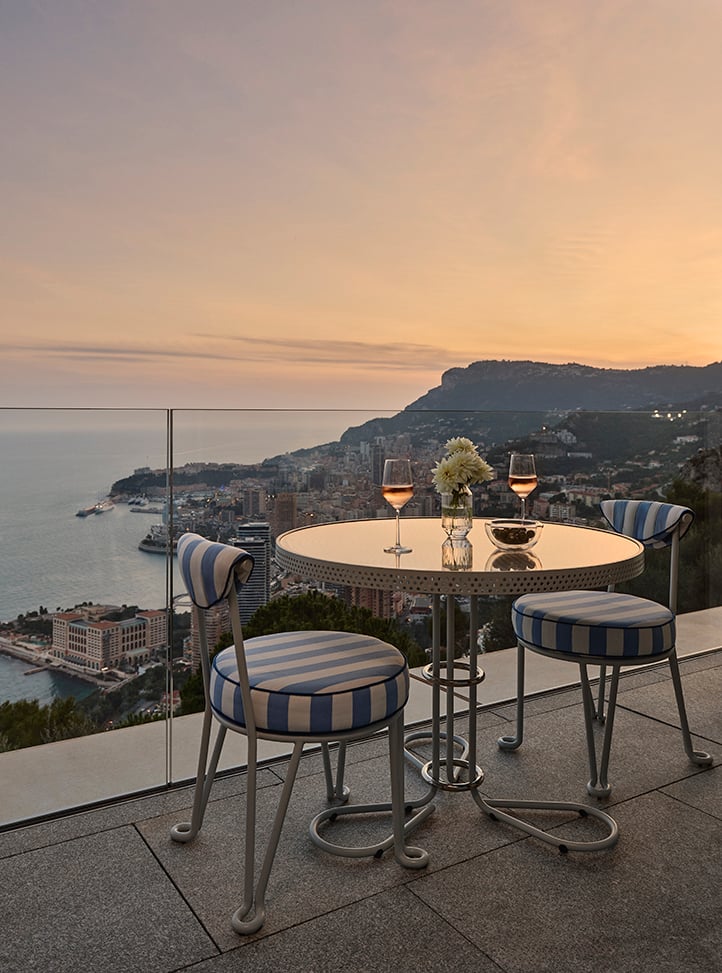 Intimate terrace setting at sunset with a round table and three blue-and-white striped chairs, overlooking the coastline and cityscape below, with two glasses of rosé wine and a small vase of white flowers on the table.
