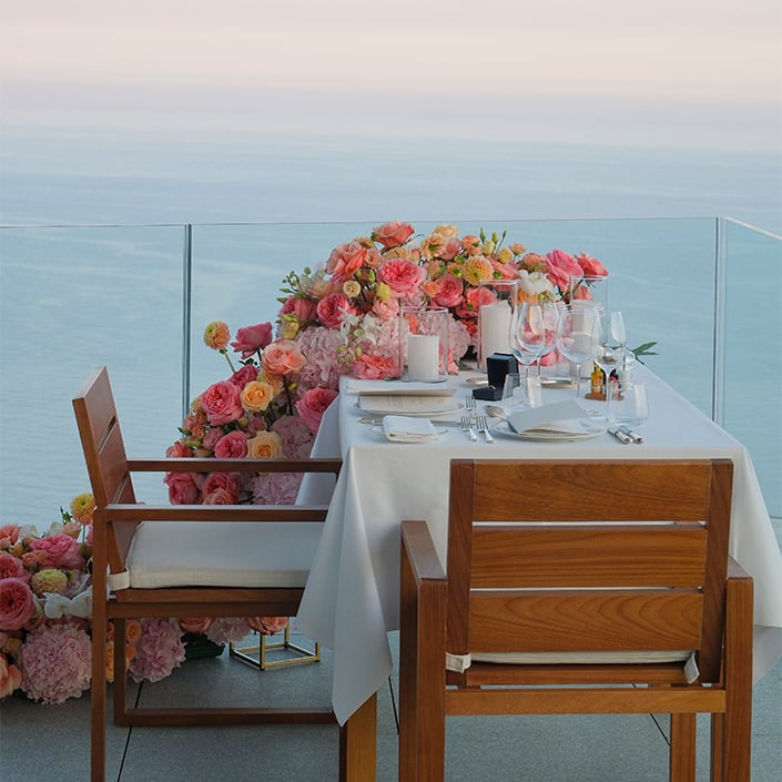A romantic oceanfront table with elegant tableware, candles, and lush pink and peach florals.