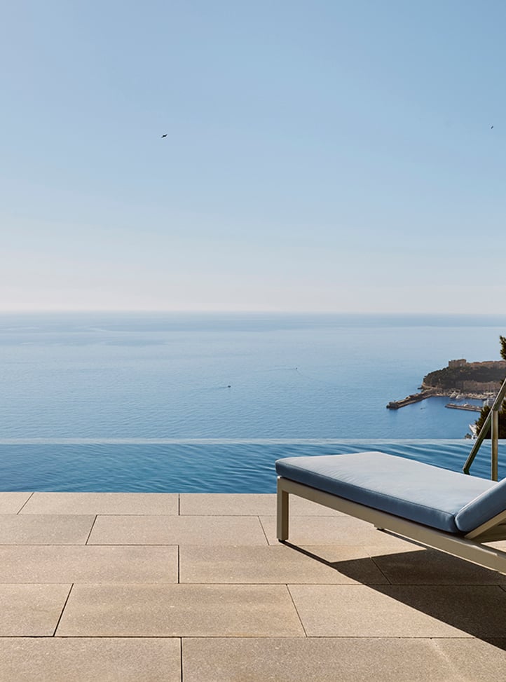 Tranquil terrace view with a single cushioned sun lounger beside an infinity pool, overlooking the calm blue sea and a distant coastline under a clear sky.