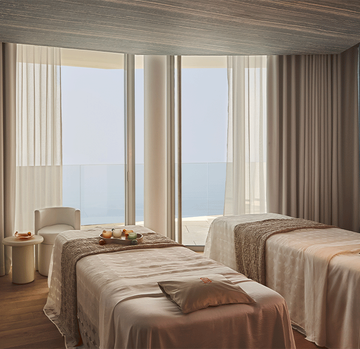 Salle de soin élégante avec deux tables de massage recouvertes de draps satinés, près de baies vitrées donnant sur la mer, voilages légers filtrant la lumière naturelle pour une ambiance apaisante.