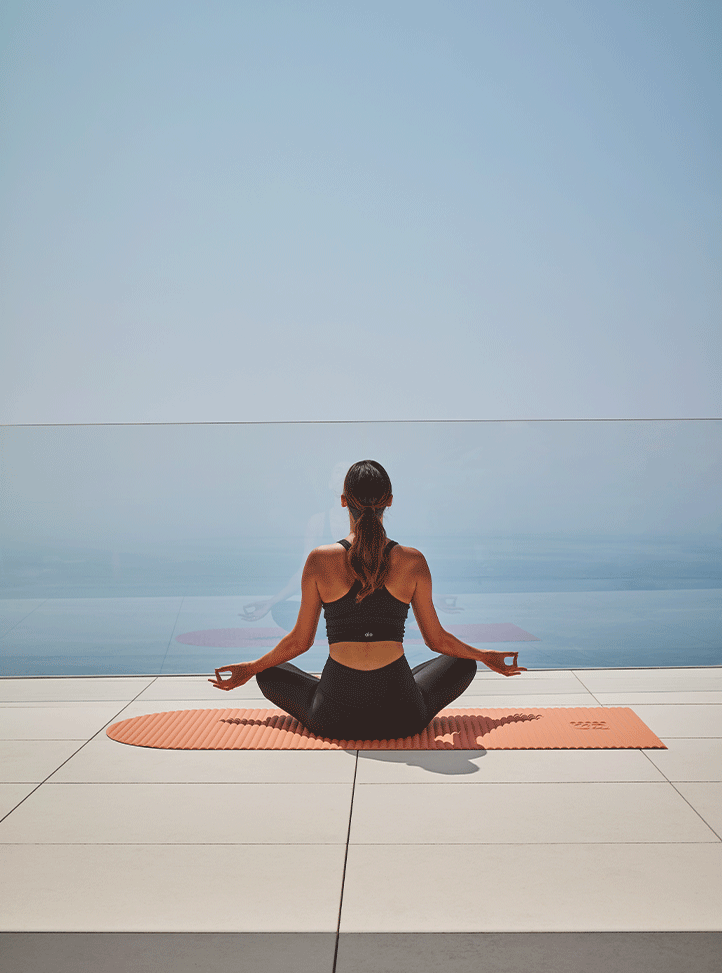 Femme en tenue de sport assise en lotus sur un tapis orange, méditant face à une vue panoramique sur la mer depuis une terrasse minimaliste aux dalles blanches.