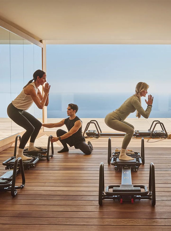 Deux hôtes pratiquent des exercices sur reformer avec un coach sur une terrasse en bois face à la mer.