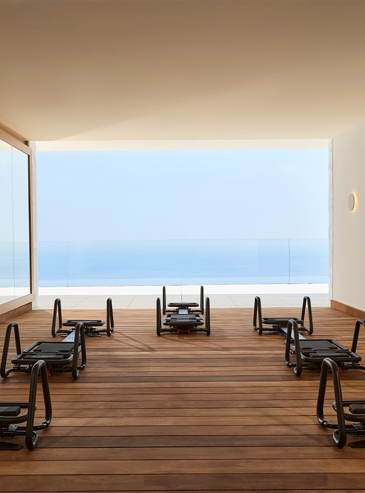 Open-air fitness studio with wooden flooring and a row of modern reformer machines arranged in a semi-circle, facing a panoramic ocean view through a frameless glass balustrade.