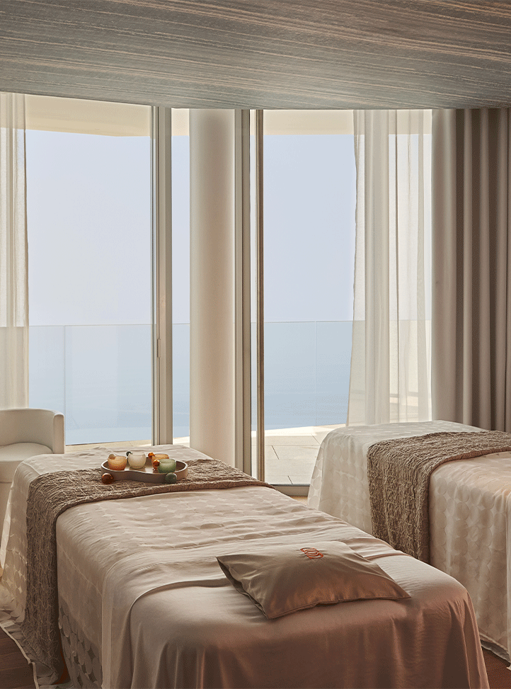 Close-up view of a tranquil spa treatment room with two massage beds covered in soft cream linens and textured throws, set beside tall windows revealing a calm sea view beyond sheer curtains.