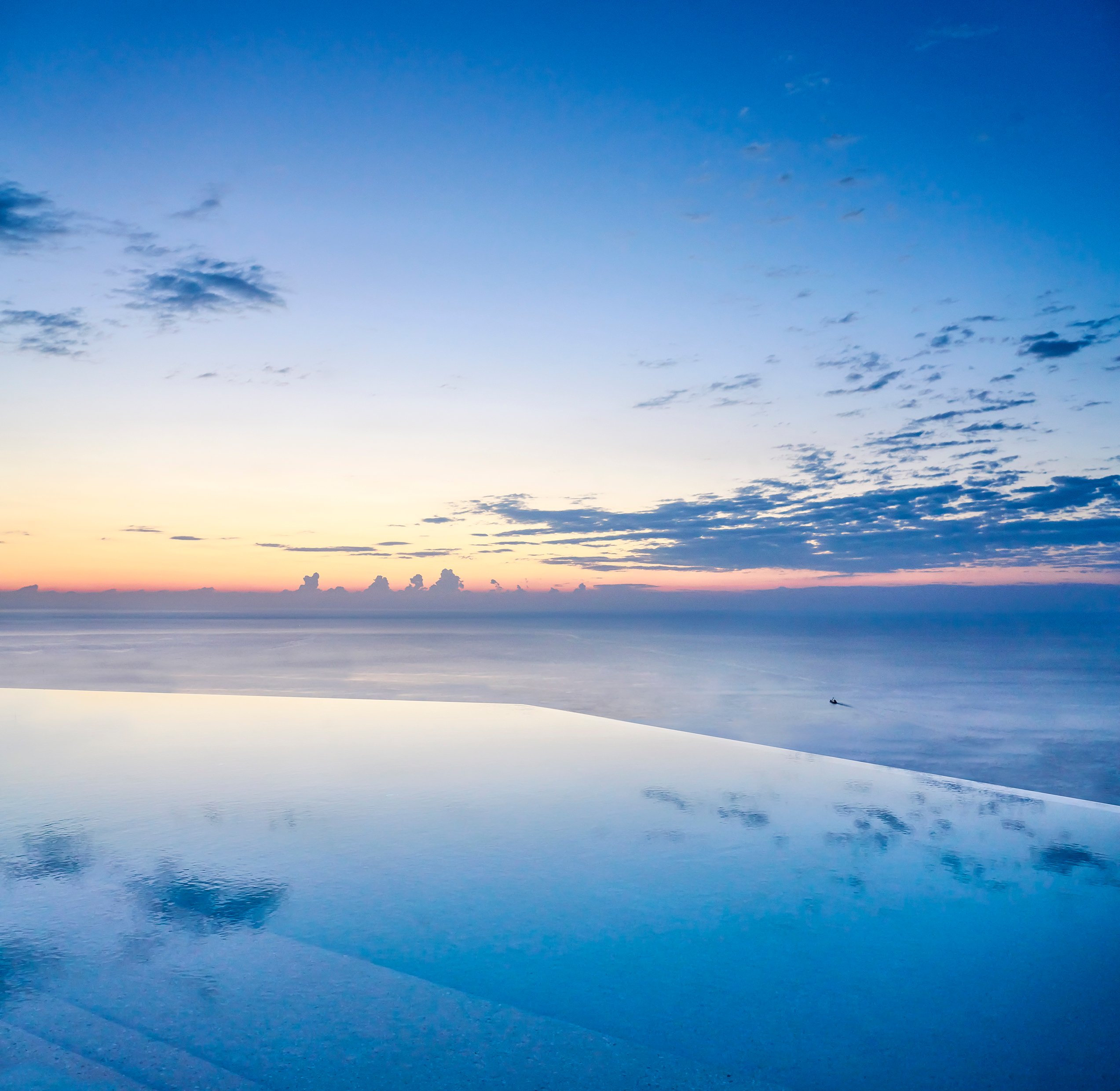 Infinity pool overlooking a calm ocean at sunrise, with soft pastel skies and scattered clouds.